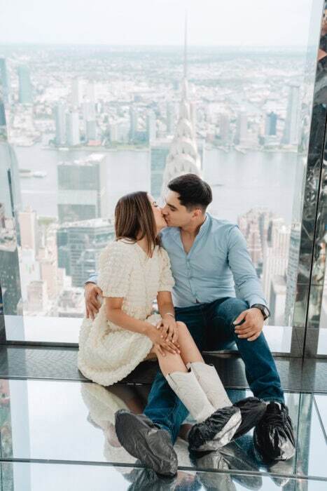 Romantic couple sharing a kiss with New York City skyline in the background from an observation deck.